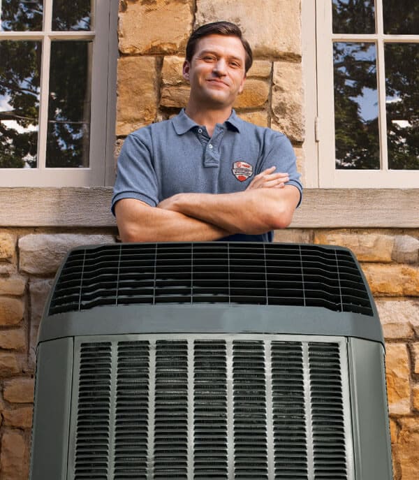 Man with crossed arms standing behind an air conditioning unit outside a building, specializing in AC Repair.