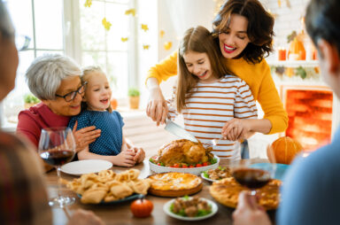 Family gathered around a table enjoying a Thanksgiving meal. A woman helps a young girl carve a roast chicken. Other dishes and glasses of wine are on the table, while the fireplace warms the room, nearly spoiling the party with its noisy crackle in the background.