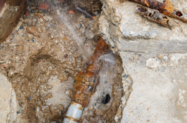 A rusty metal pipe, reminiscent of the evolution of plumbing, is leaking water onto a muddy surface inside a concrete trench.